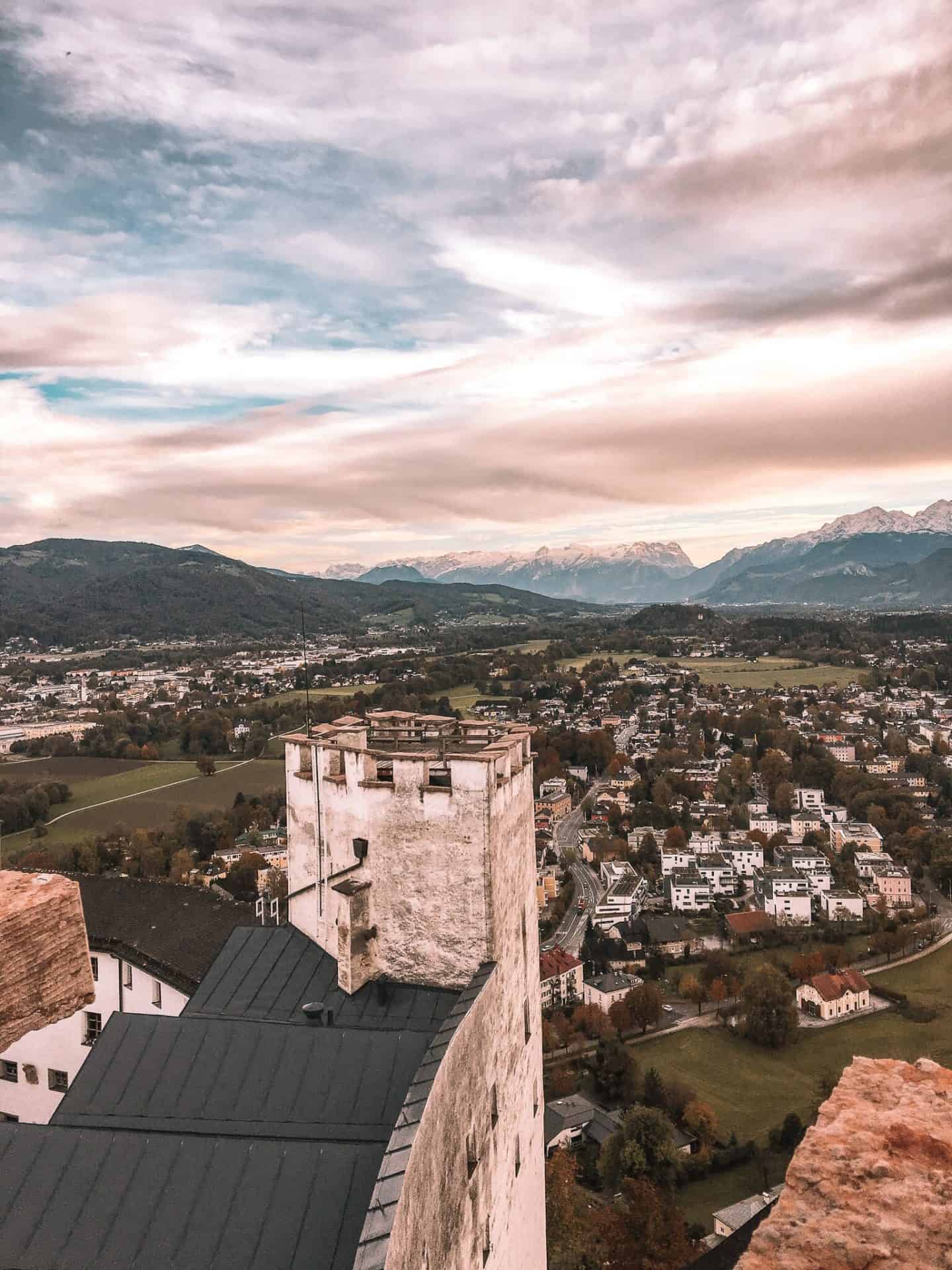 View from Hohensalzburg Fortress overlooking Salzburg, Austria, with the city’s rooftops, green fields, and distant mountains under a dramatic cloudy sky.