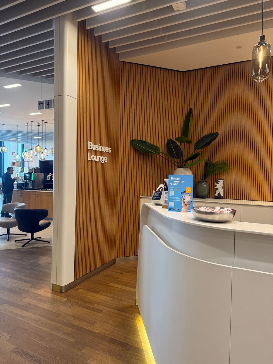 Entrance to an airport Business Lounge with a sleek white reception desk, warm wood paneling, and seating area in the background, creating a calm atmosphere for travelers.