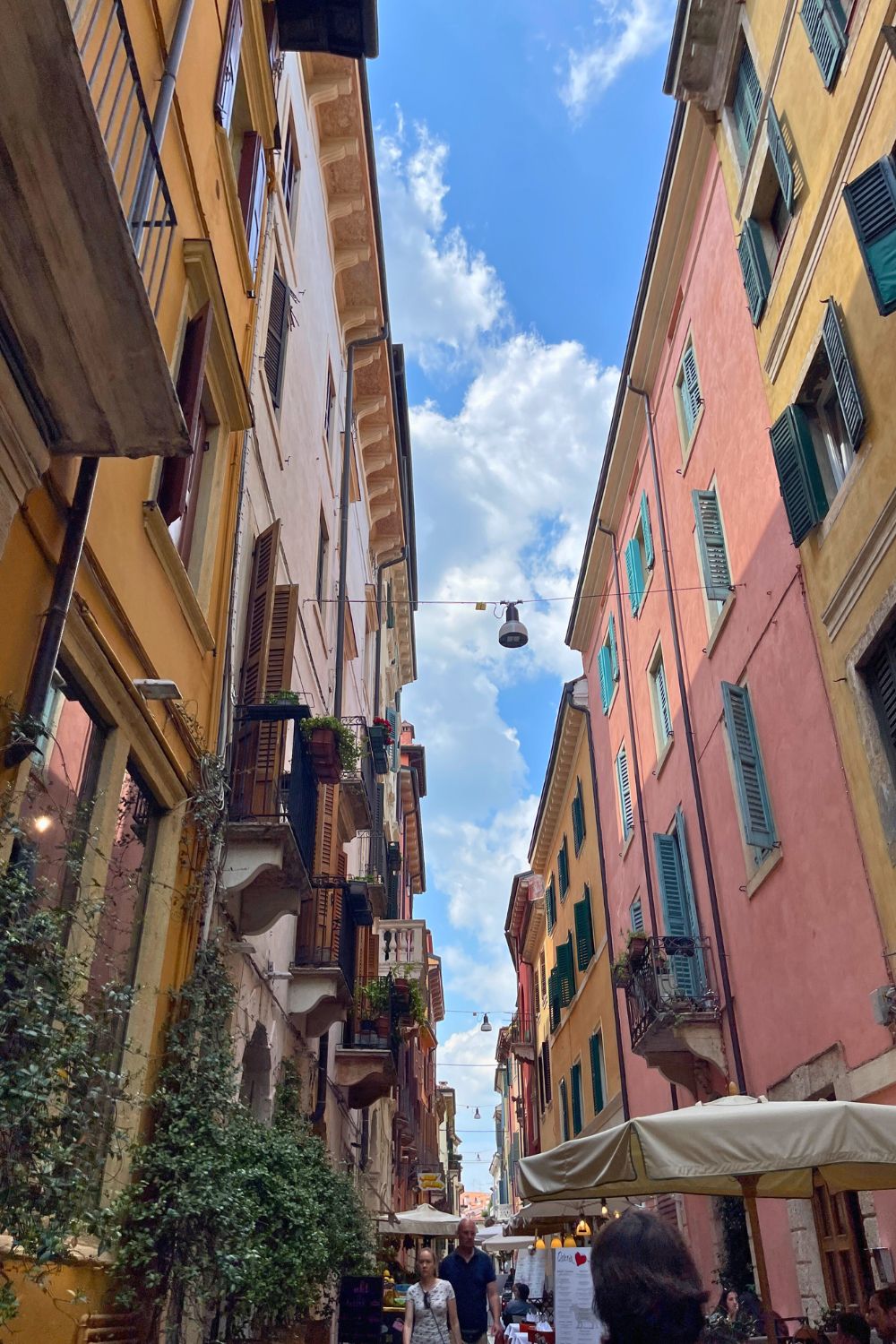 A view of a narrow street in Verona, Italy, lined with tall, colorful buildings with shutters and balconies. The sky is bright blue with fluffy white clouds, and the street is adorned with greenery and people walking below.