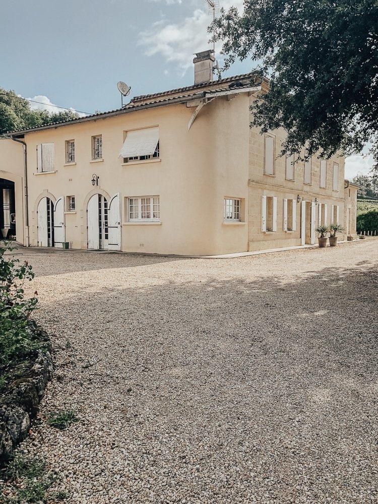 A stately cream-colored building with classic French windows and doors, surrounded by a pebbled courtyard and lush greenery, typical of the elegant estates found on the best Saint-Emilion wine tours.