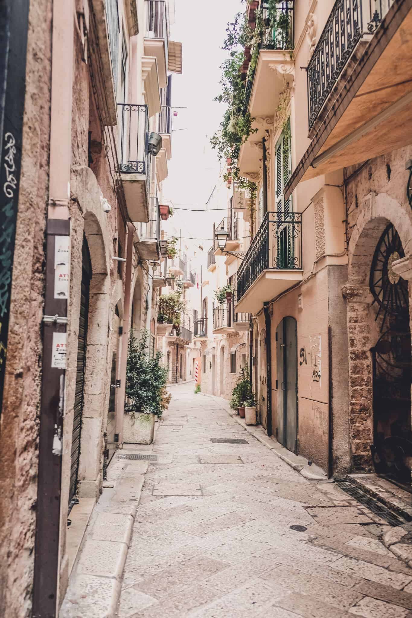 A narrow, picturesque alley in Bari, Italy, lined with old stone buildings and balconies adorned with plants. The alley is paved with stone and has a charming, quaint atmosphere.