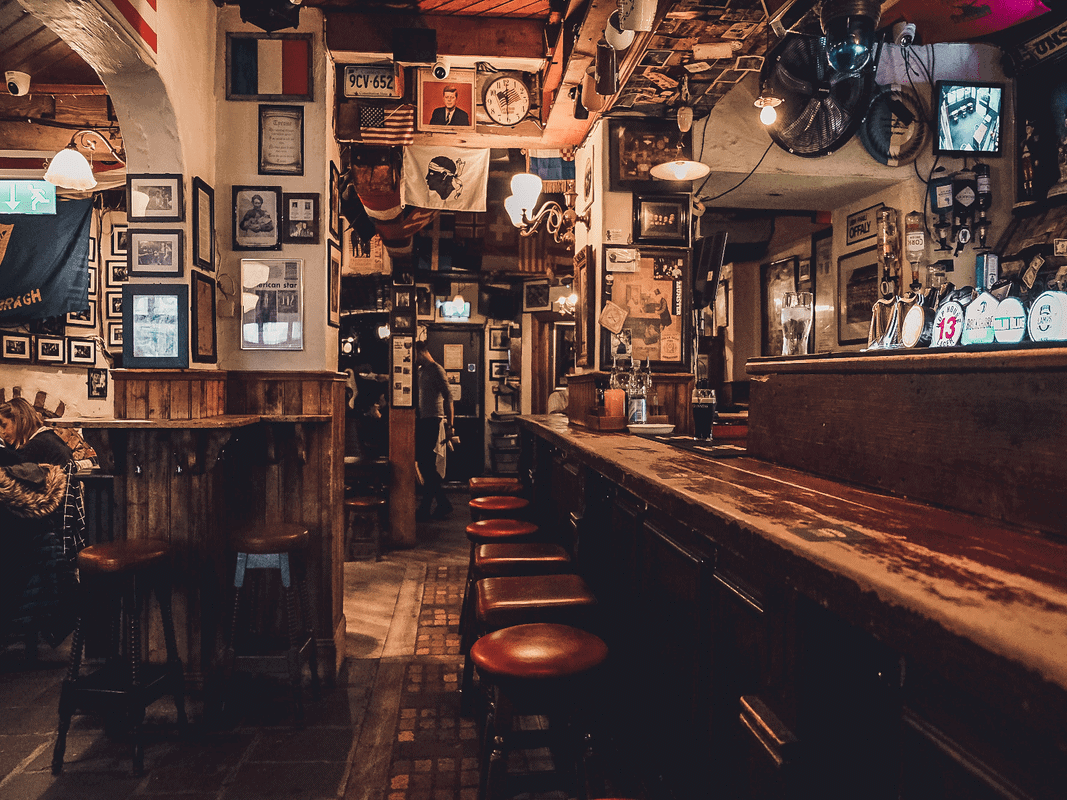 A cozy traditional Irish pub in Dublin, with a warm atmosphere, decorated with historical memorabilia and framed pictures, and a line of stools at the wooden bar waiting for solo travelers to take a seat.