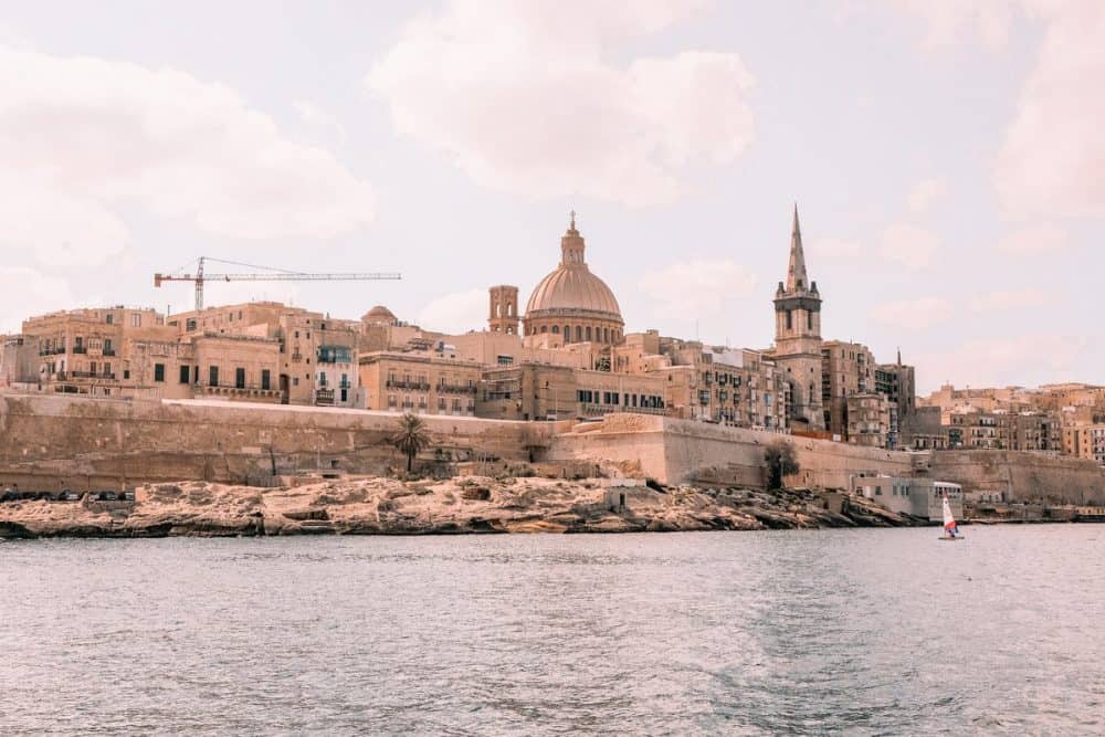 Valletta's iconic skyline, with its historic sandstone buildings and the majestic dome of St. Paul's Pro-Cathedral, overlooks the clear blue waters of the Mediterranean Sea with a windsurfer in the foreground.