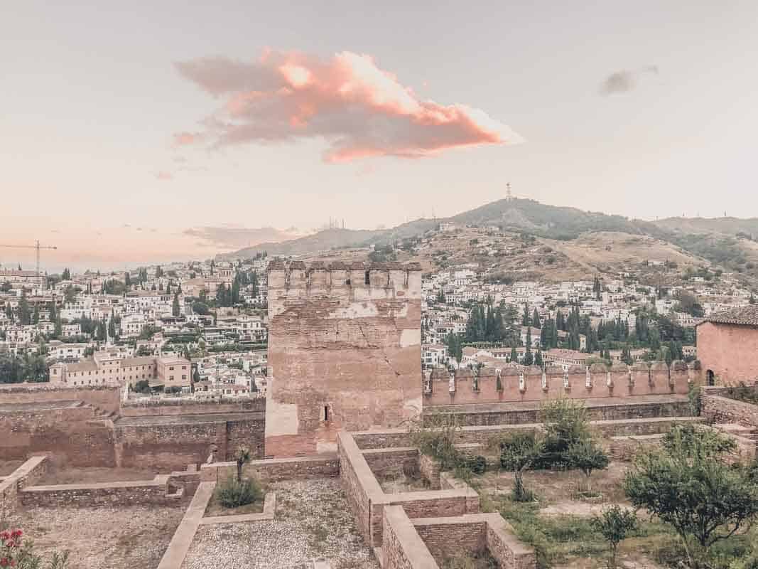 The image shows a scenic view of the historic city of Granada, Spain, with the Alhambra's fortress walls in the foreground. The city spreads across the valley and into the distant hills, with whitewashed buildings dotting the landscape. The sky is soft and pink, illuminated by a glowing cloud at sunset or sunrise, adding a serene atmosphere to the scene.






