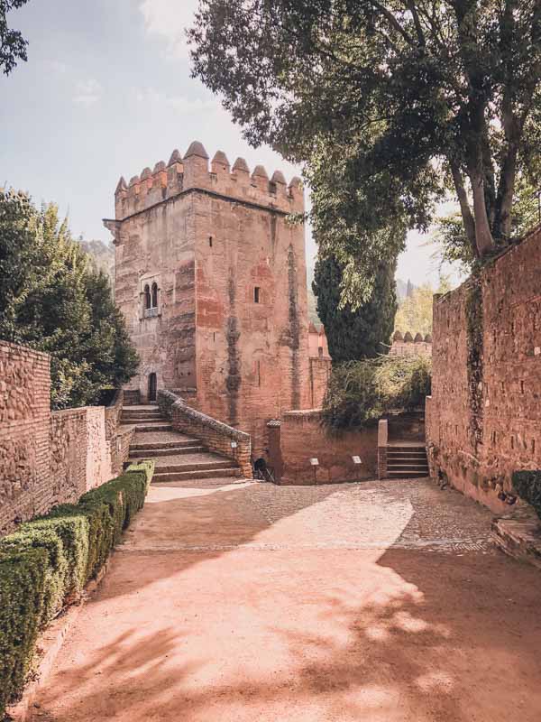 Historic stone tower with crenelated battlements, surrounded by high walls and lush greenery. The scene is bathed in soft sunlight, casting shadows on the cobblestone path that leads up to the structure. Trees and manicured bushes frame the path, adding a touch of nature to the medieval architecture.