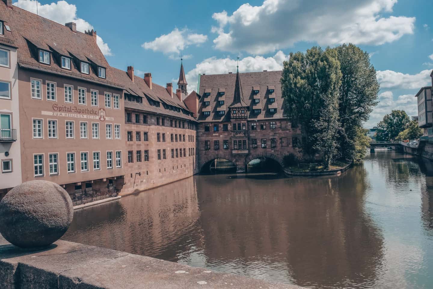 "A serene view of the Pegnitz River flowing through Nuremberg, flanked by historic sandstone buildings with steep roofs and rows of windows reflecting on the water's surface. The famous Heilig-Geist-Spital, a hospital from medieval times, stands prominent with its picturesque bridge connecting the structures, offering a glimpse into the city's storied past on a peaceful day.