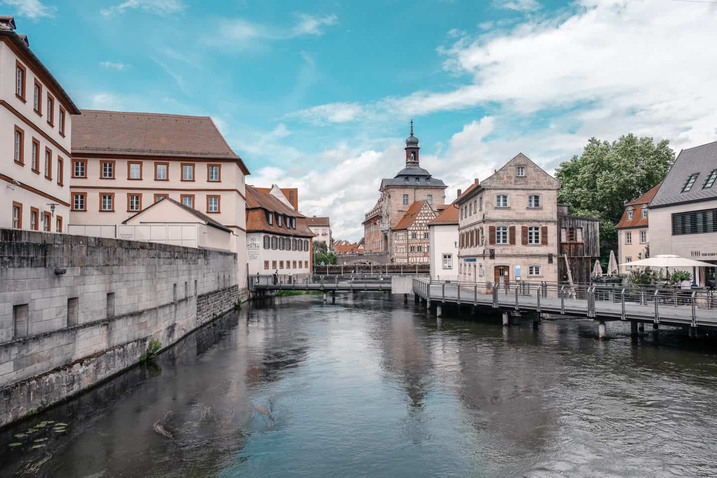 A serene river scene in Bamberg, with a view of historical buildings reflecting the city's architectural heritage. The tranquil waters of the river are bordered by stone walls, and a pedestrian bridge connects both sides, leading to charming buildings with classic Franconian half-timbered designs. The cloudy sky adds a peaceful overture to this picturesque setting.