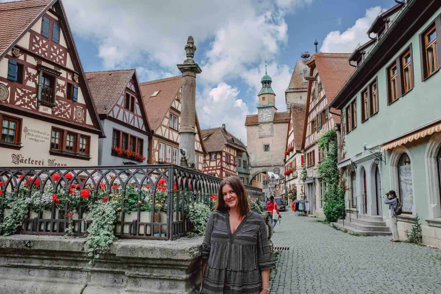 A woman stands smiling on a cobblestone street in Rothenburg ob der Tauber, with half-timbered buildings adorned with red flowers in the background. A historical clock tower rises in the distance, adding to the old-world charm of the scene.