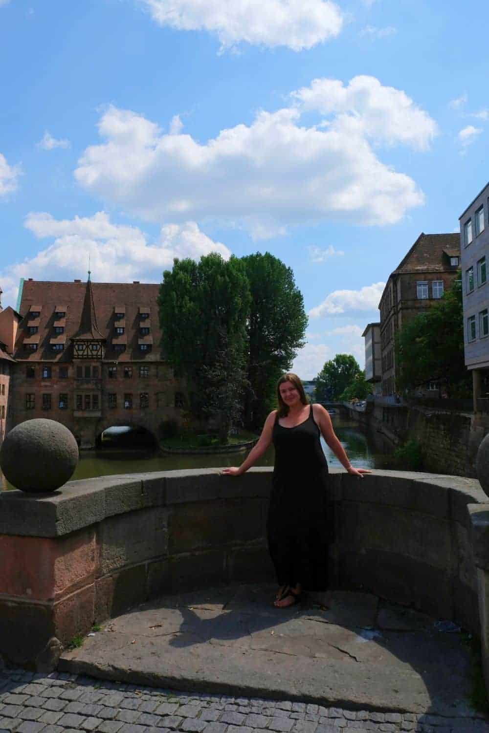 The same woman smiles as she leans on the balustrade of a bridge in Nuremberg on a sunny day, with traditional German architecture and lush trees behind her, embodying the joy of exploring the city's rich history and culture.