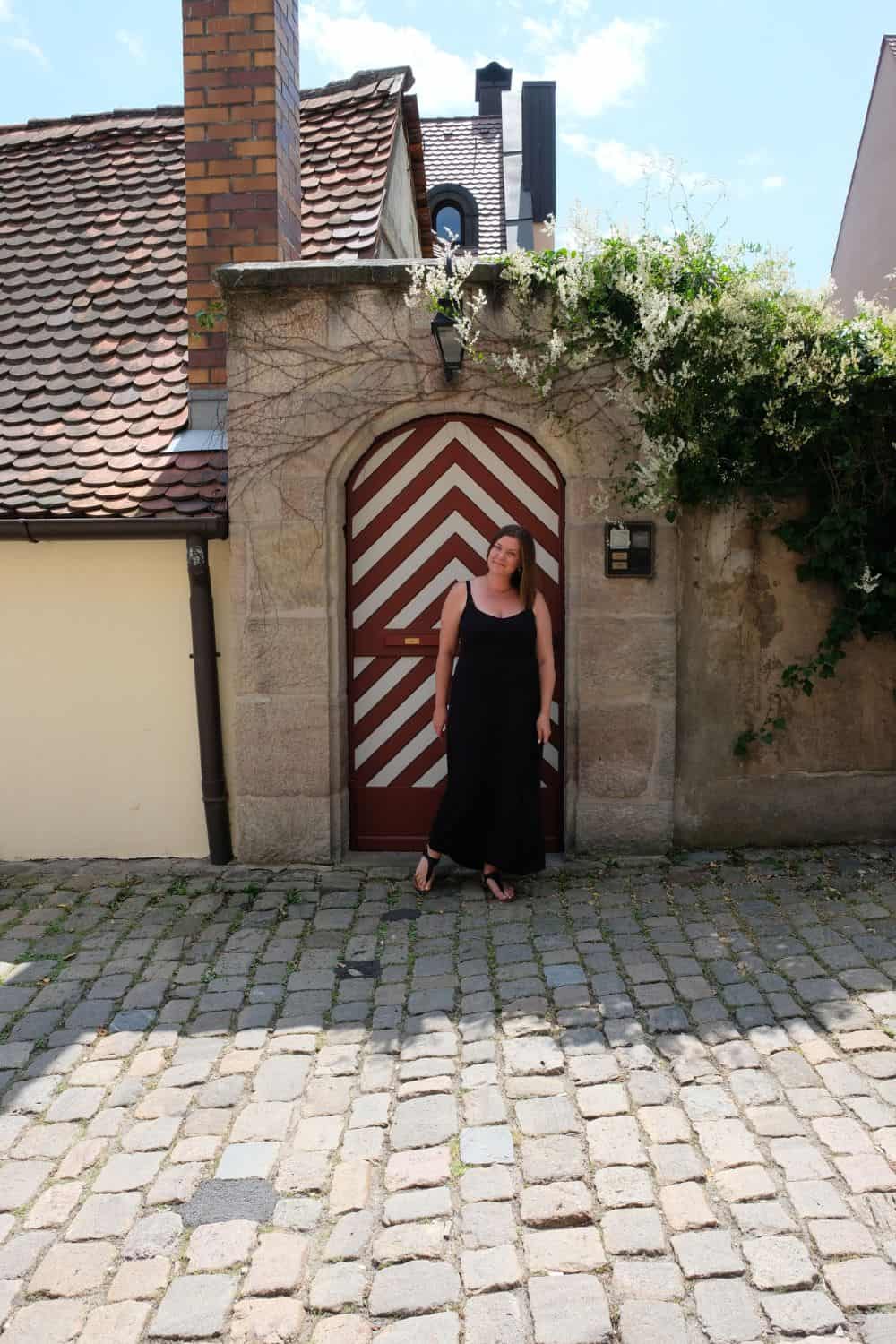 A woman in a black dress stands smiling by a distinctive red and white patterned door in Nuremberg, surrounded by the city's charming cobblestone streets. A cascade of white blossoms hangs over the stone archway, adding a touch of natural beauty to this quaint, historic setting, perfect for a leisurely exploration on a sunny day.
