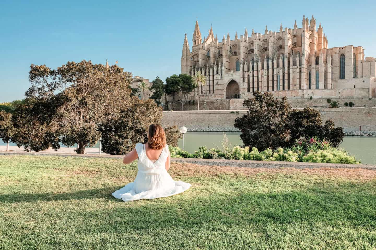An independent traveler enjoys a peaceful moment in Mallorca, sitting on the green grass, gazing at the majestic Cathedral of Santa Maria in the distance, symbolizing the beauty of solo travel.