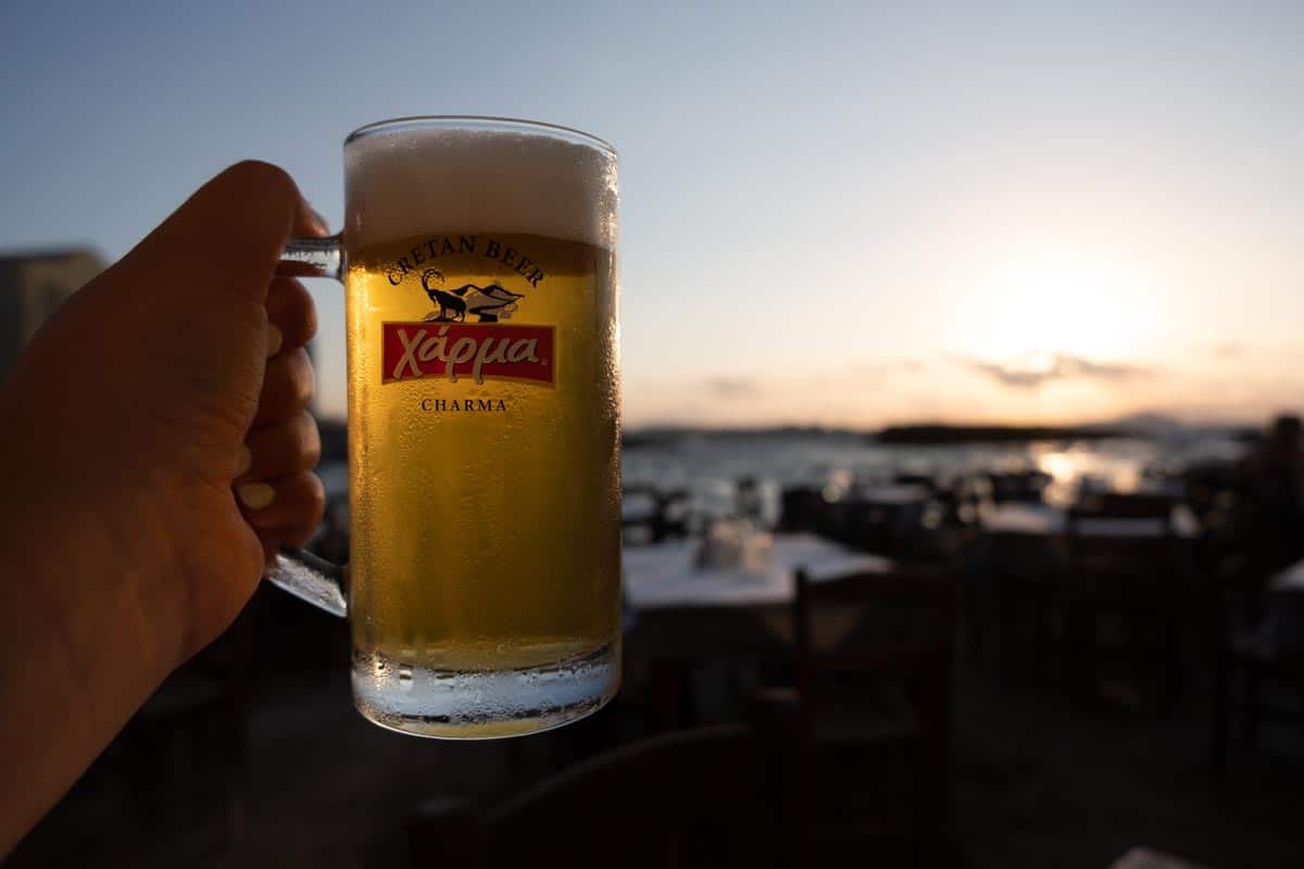 A hand holding up a beer in Crete Greece with the sunset and ocean in the background