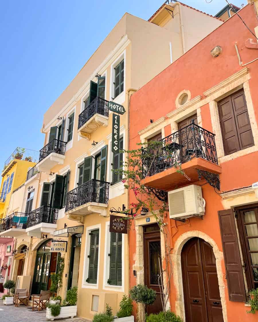 A charming street view on a warm Greek island in May, highlighting the vibrant architecture with a pastel yellow building adjacent to a salmon-colored structure, both adorned with black iron balconies and potted plants, under a clear blue sky.