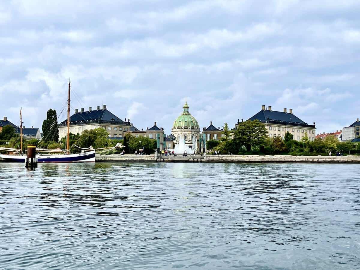 A view across the calm waters of a Copenhagen canal, showcasing the elegant Frederik's Church with its distinctive green dome and baroque architecture, commonly known as the Marble Church. The church is flanked by classic European buildings under a partly cloudy sky. In the foreground, a classic wooden sailboat is moored along the quay, adding a touch of maritime charm to the urban landscape. The water reflects the sky and structures, creating a tranquil and picturesque scene.