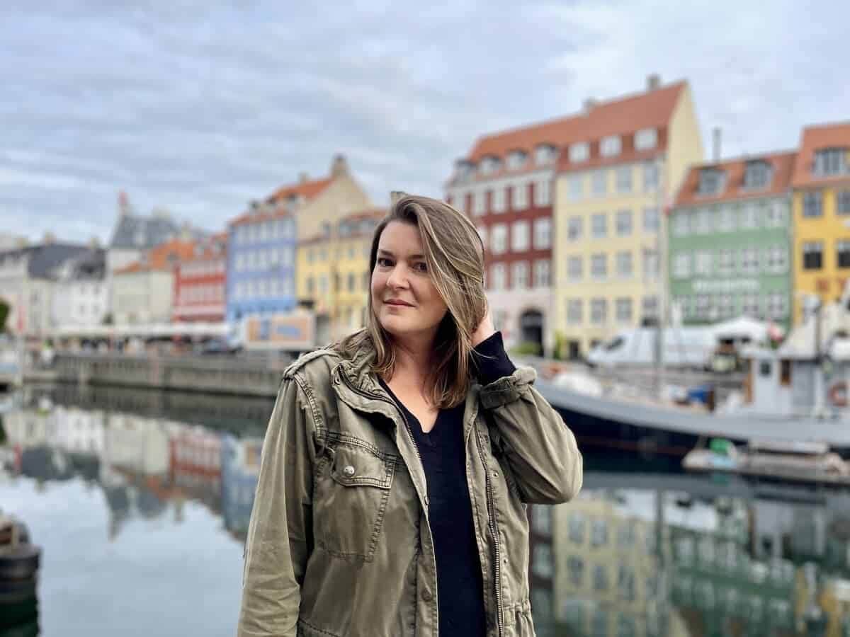 A solo female traveler stands before the iconic Nyhavn canal in Copenhagen, Denmark. She's positioned on the left side of the frame, smiling gently while touching her hair, and wearing a casual outfit with a green jacket over a black top. Colorful historic houses line the background, reflecting on the still water along with moored boats, under a soft, overcast sky. The image conveys a sense of relaxed exploration, typical of a solo travel experience.