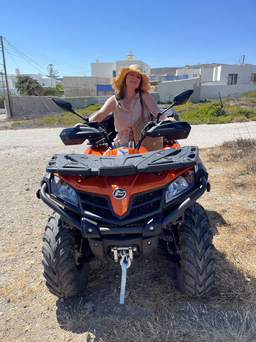 A woman posing on an orange ATV with a backdrop of traditional Greek architecture in Milos.