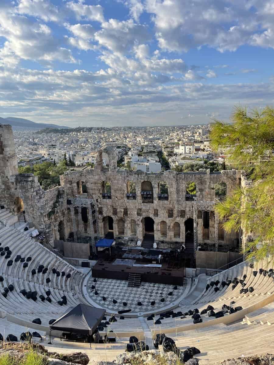 Ancient grandeur meets modern day in the Odeon of Herodes Atticus in Athens, where the timeless stone theater overlooks a sprawling cityscape, under a sky punctuated by wispy clouds, ready to host a cultural performance.