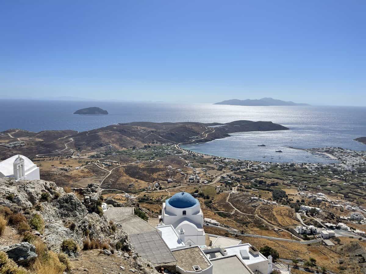 Alt text: "Elevated view over the rugged terrain of a Greek island, with a panoramic vista of the sparkling Aegean Sea. A traditional white church with a blue dome sits in the foreground, overlooking a valley dotted with buildings, winding roads, and sparse vegetation.