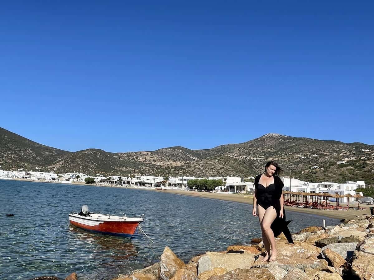 A woman in a black swimsuit enjoying a solo trip on the rocky shore of a Greek island, with a small red boat floating in the clear blue waters and white buildings nestled against the hills in the background.