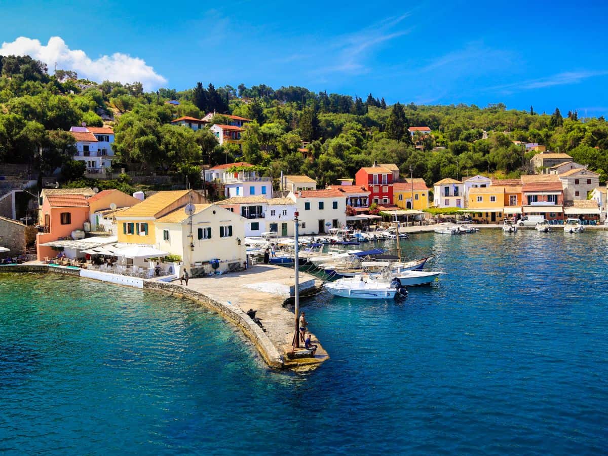 A picturesque view of a charming Greek island harbor lined with colorful houses. Lush greenery adorns the hills above, while leisure boats docked in the clear blue water invite exploration. A perfect snapshot of a serene solo travel destination in the Greek islands.