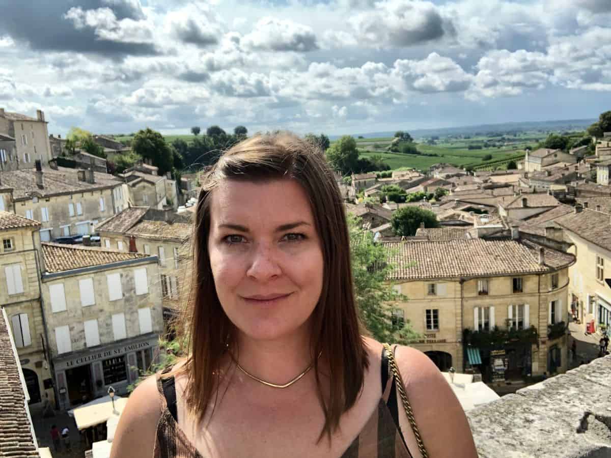A woman stands in the foreground, a smile beginning to form, with the ancient rooftops of Saint-Emilion stretching out behind her under a dramatic sky, capturing the essence of a solo traveler's experience on in best Saint-Emilion.