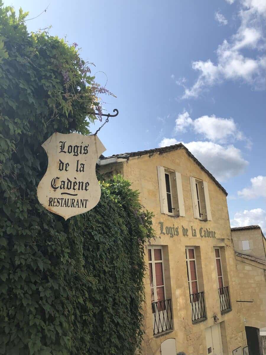 A charming, ivy-draped sign reading 'Logis de la Cadène' points to the restaurant below, set in a classic yellow stone building under a sunny blue sky in Saint-Emilion, inviting guests on wine tours to savor local gastronomy.