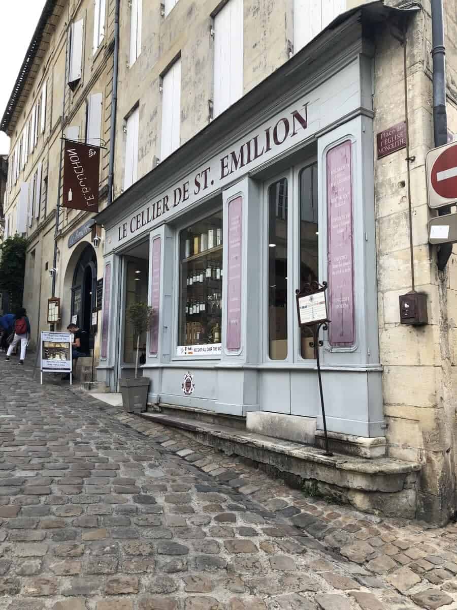 The quaint façade of 'Le Cellier de St. Emilion' wine shop on a cobbled street in downtown Saint-Emilion, with its weathered shutters and classic signage, inviting passersby to explore local wine selections.