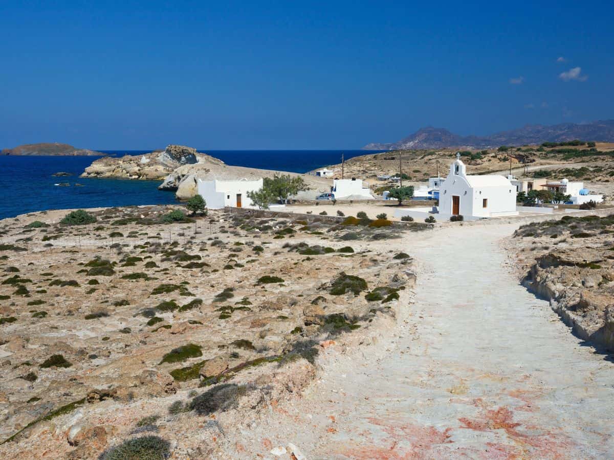 A dirt road in Milos with a white church and ocean in the background.