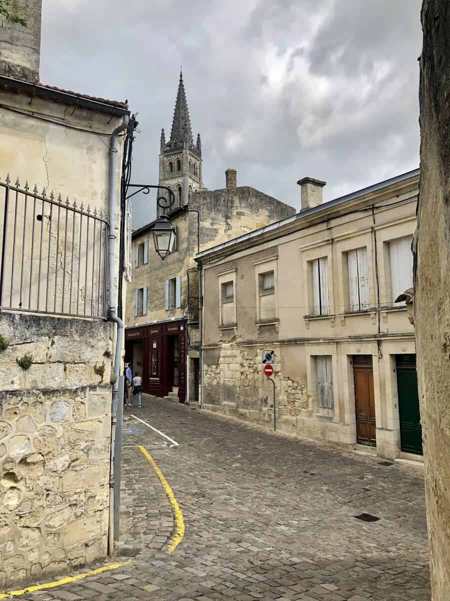 A cobblestone street in Saint-Emilion meanders between aged stone buildings leading towards an imposing church spire under a dramatic cloudy sky, a view characteristic of the region's storied wine tours.