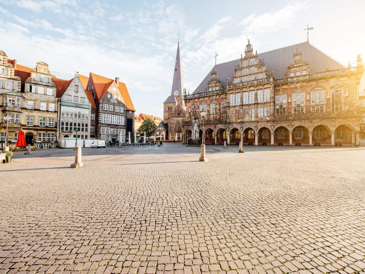 A serene morning view of the historic Market Square in Bremen, Germany. Sunlight bathes the scene, highlighting the intricate facades of traditional German buildings with their distinct stepped gables and the ornate Renaissance architecture of the Town Hall.
