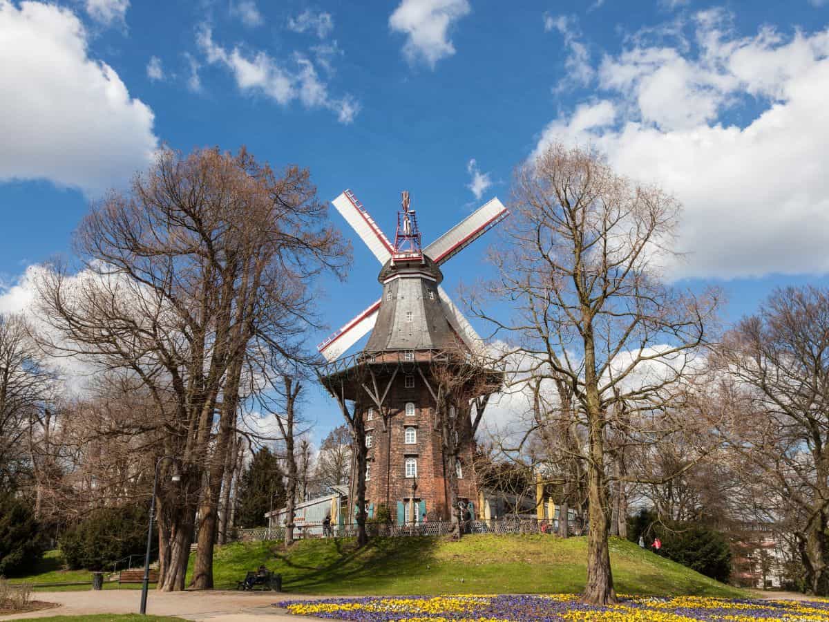 A traditional windmill set against a partly cloudy sky in Bremen, Germany. The windmill's red and white sails are prominent, and it stands atop a small hill, surrounded by leafless trees suggesting an early spring or late autumn season.