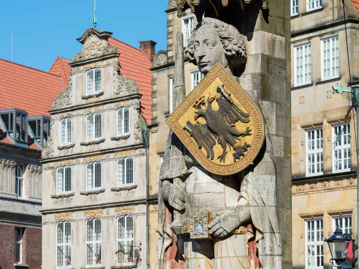 A close-up of a stone sculpture featuring a classical face, possibly a part of a larger monument or fountain, holding a shield emblazoned with the Bremen coat of arms, which displays a black eagle on a golden background.