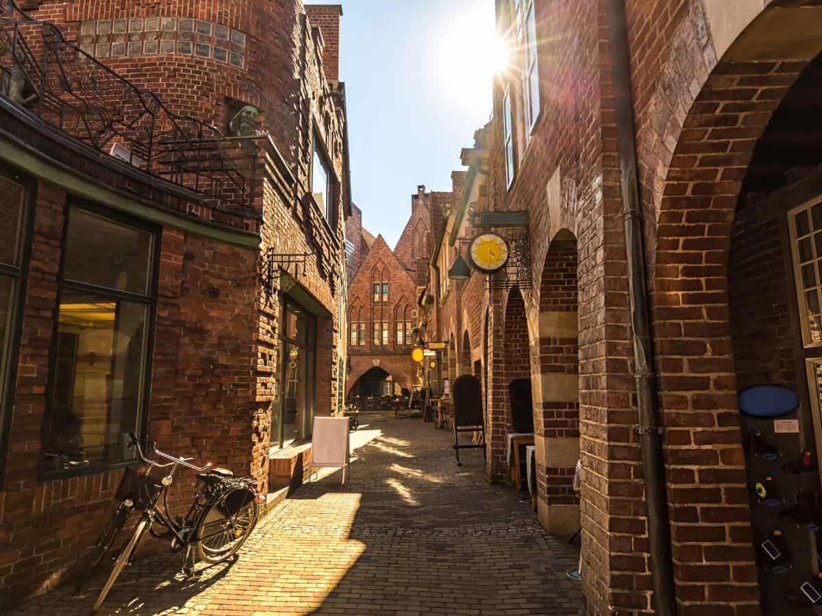 A narrow alley in Bremen, Germany, paved with bricks and flanked by historical brick buildings with a distinctive Hanseatic architecture. On the left, a bicycle is parked against a window beneath ornate wrought-iron railings. On the right, the buildings feature arched doorways, with a prominent clock hanging from a wrought-iron bracket.