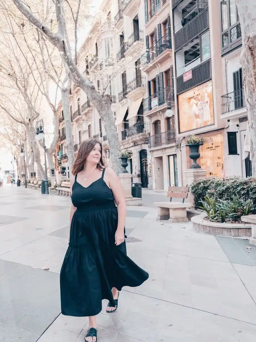 A confident solo explorer walks down a picturesque street in Mallorca, her casual elegance matching the relaxed vibe of the tree-lined boulevard, perfect for a solo travel adventure