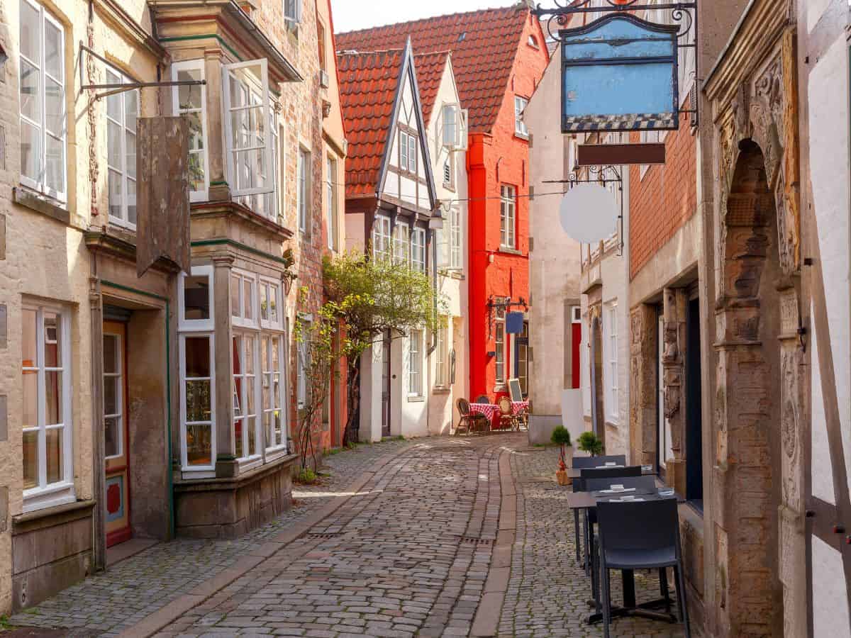 A cobblestone street in Bremen, lined with charming, colorful, old European buildings with gabled roofs. The architecture suggests a blend of residential and commercial properties. To the left, there's a building with large windows, and an empty signboard hanging above, indicating a place for a shop or a cafe. To the right, a vibrant red building stands out, next to a white one with an arched doorway. Outdoor furniture suggests a quaint café setting.