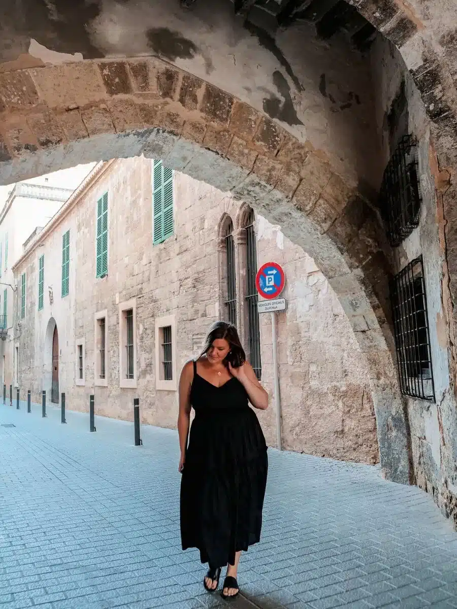 A woman in a flowing black dress walks alone down a cobblestone street in a historic European city. She looks down, a soft smile on her face, as she passes under an ancient stone archway that frames the scene. 