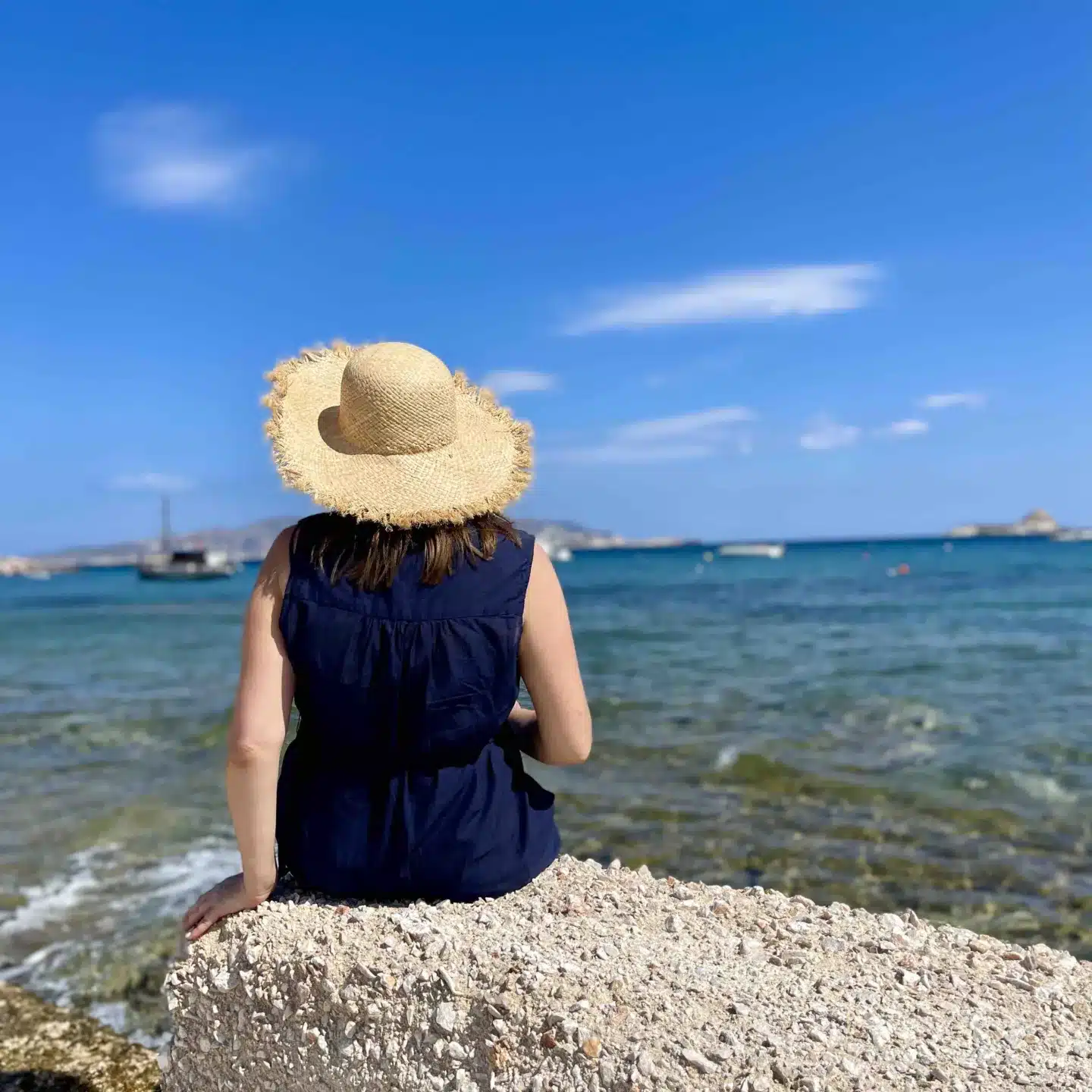 A woman in a blue dress and straw hat sitting on a rock in front of the ocean in Milos.