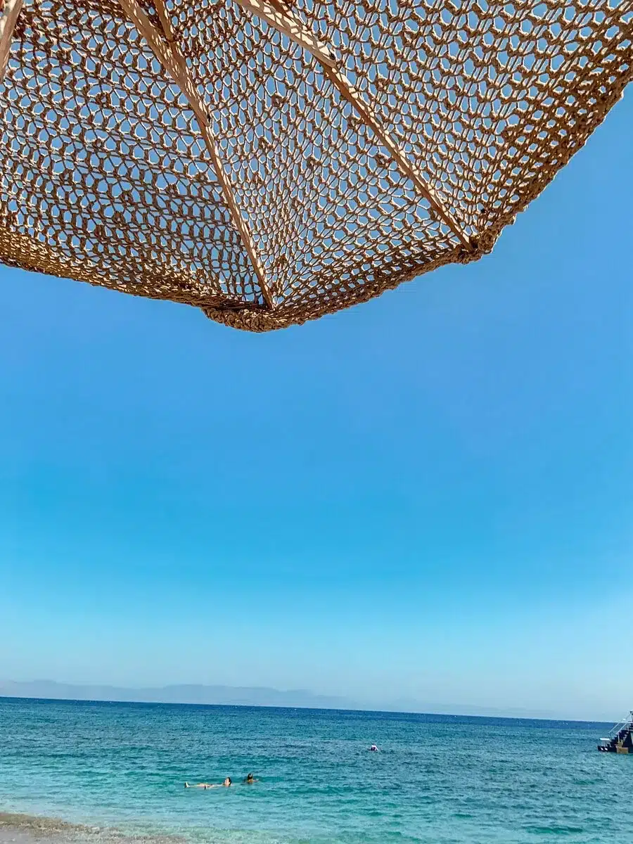 A serene beach view framed by a straw sunshade, looking out over the clear, calm waters of the Aegean Sea at the Greek island of Rhodes, renowned as one of the hottest Greek islands for its sunny weather and picturesque landscapes.