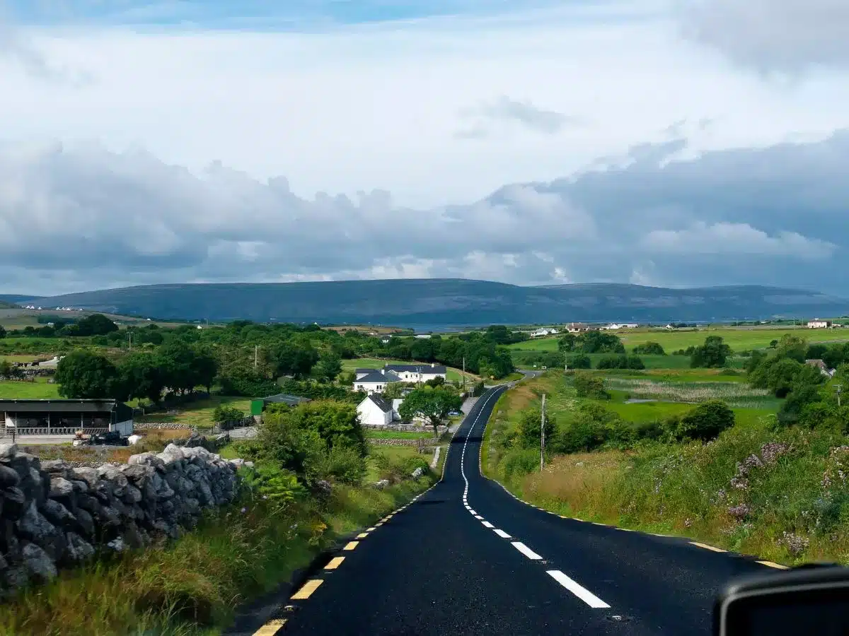 View from the driver's perspective while renting a car in Northern Ireland, showcasing a winding road leading through a picturesque rural landscape with lush green fields, stone fences, and distant mountains under a partly cloudy sky.