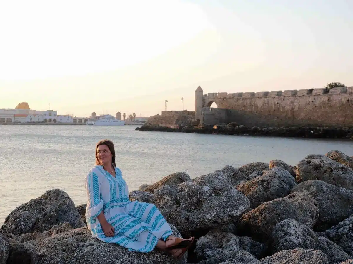 A woman sitting on the rock with the ocean and historical castle walls in the background.