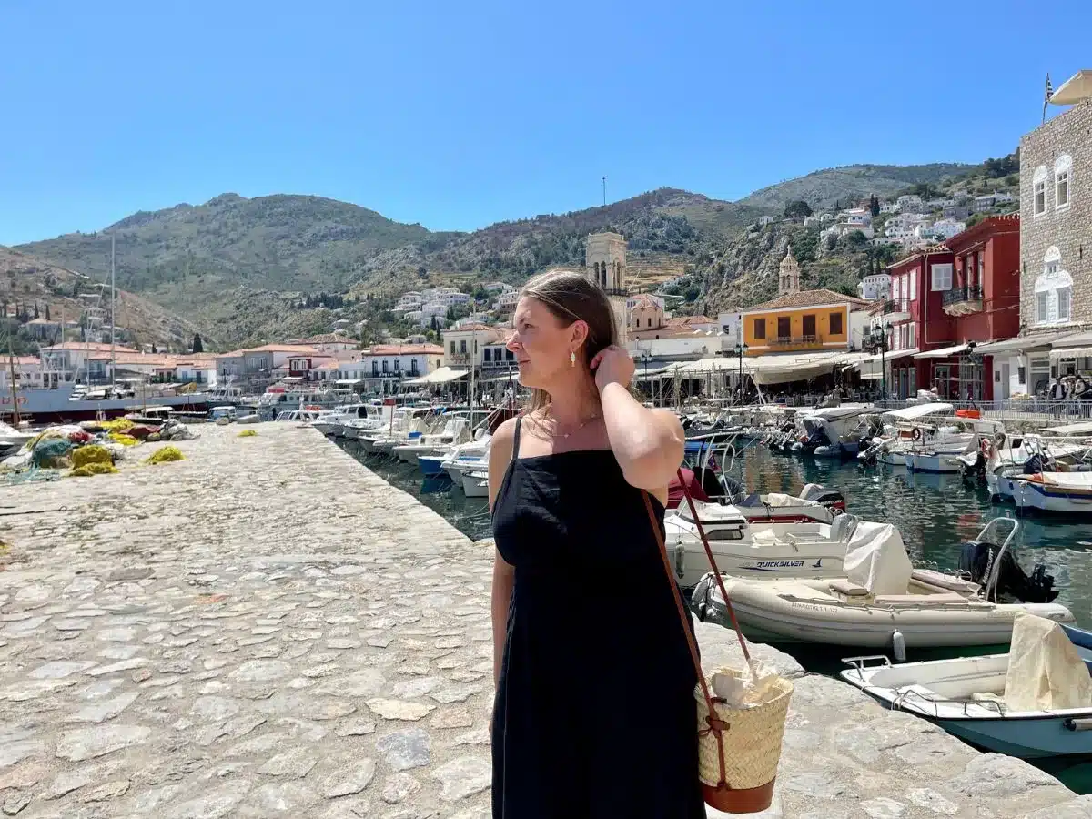 A woman in a simple, elegant black dress stands by a seaside promenade, her gaze thoughtfully directed towards the horizon. She's holding her hair back, suggesting a gentle breeze, with a straw bag casually slung over her shoulder. The Mediterranean setting, with boats moored in the harbor and historic buildings along the waterfront under a bright blue sky, reflects a sense of discovery and solitude that can come with traveling alone.