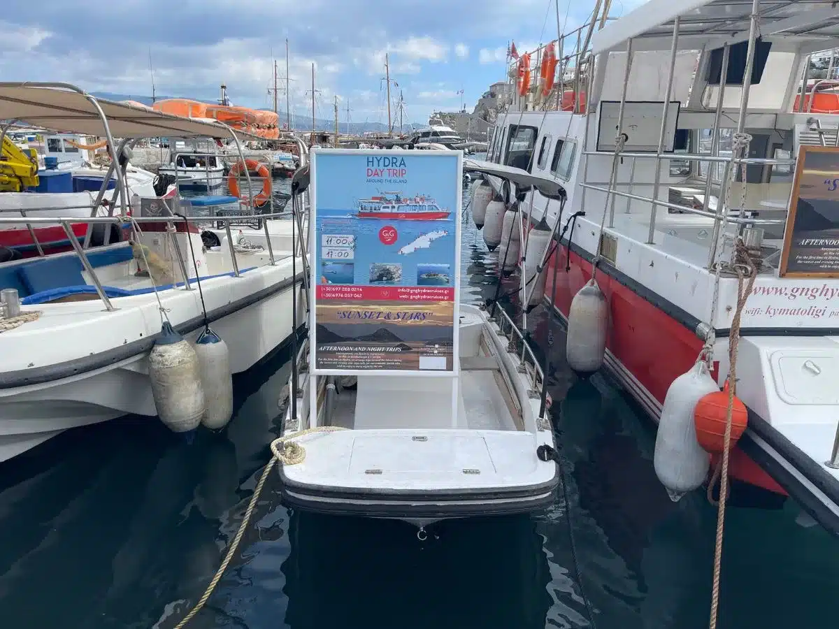 Advertisement for 'HYDRA DAY TRIP' tours displayed on a signboard at Hydra port, surrounded by moored boats, offering experiences like 'SUNSET & STARS' to explore the island's beauty.