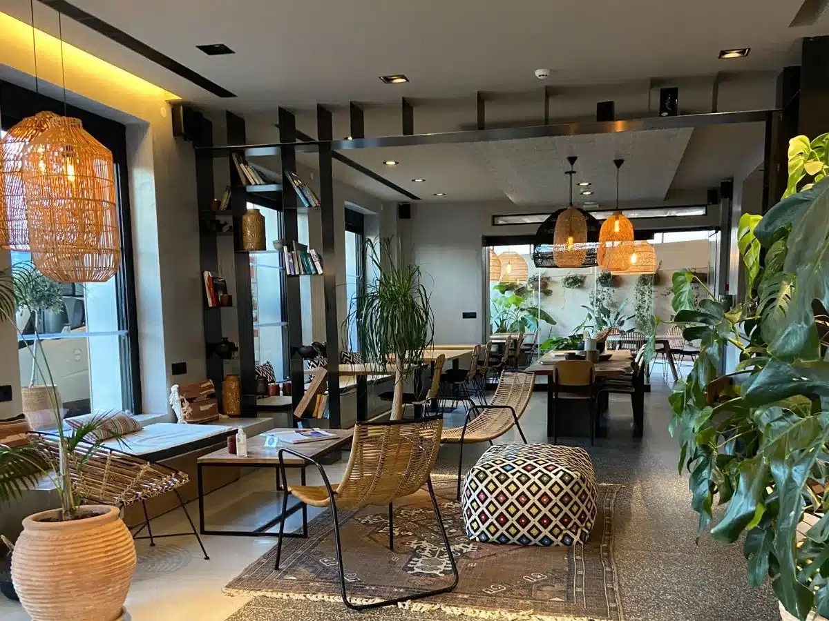 The welcoming lobby of Cook's Club Rhodes is adorned with rattan pendant lighting, woven furniture, and lush indoor plants, creating a stylish and relaxed atmosphere for guests to enjoy.
