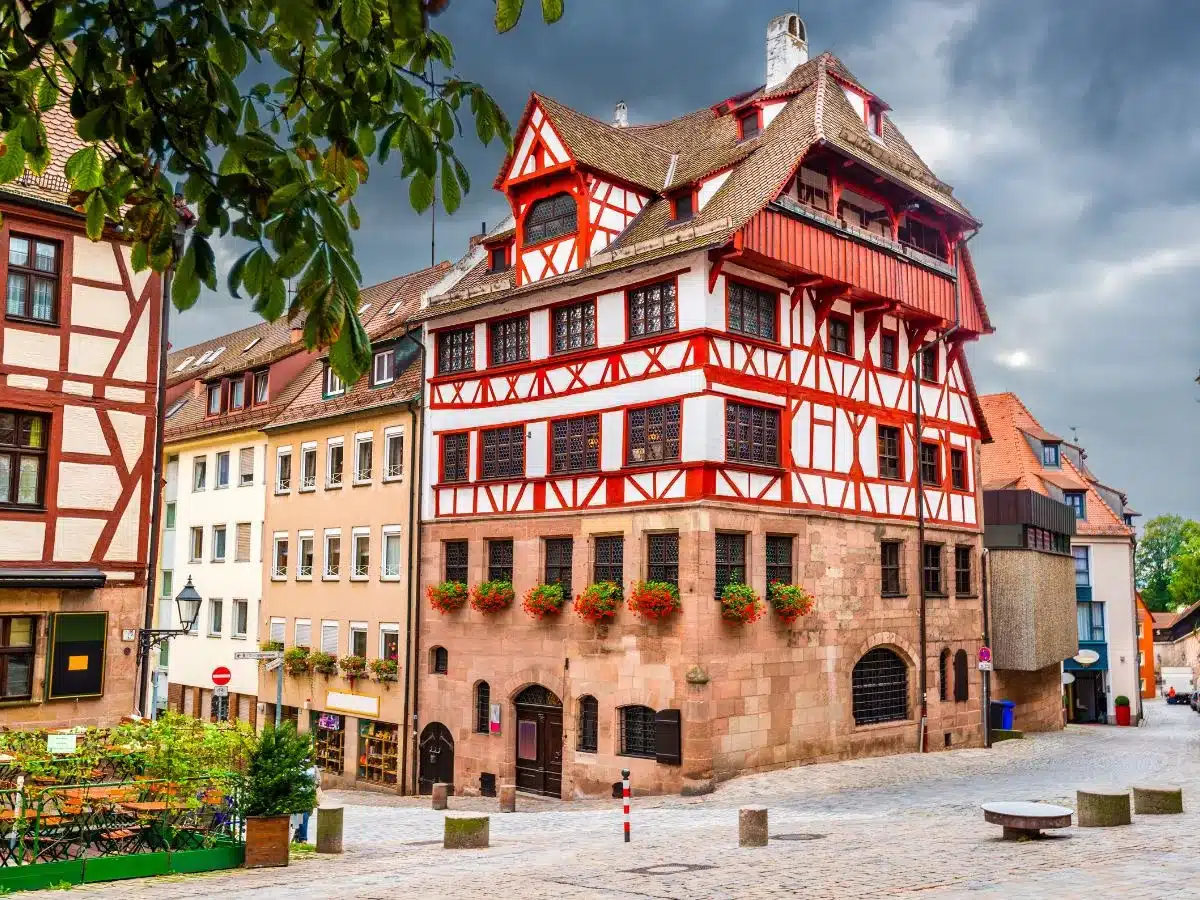 A picturesque cobblestone square at Albrecht Dürer's House. Dominated by a striking half-timbered building with bright red beams and flower boxes. The building's traditional Franconian architecture, complete with a peaked roof and detailed facades, is a testament to the city's rich heritage and provides a charming atmosphere for passersby. 