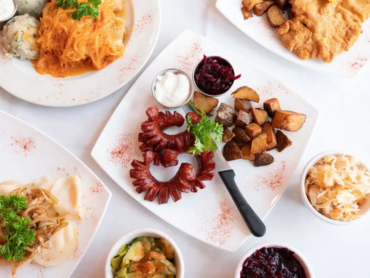 A top-down view of a variety of traditional German dishes, showcasing a colorful assortment on white plates. The spread includes sausages arranged in a circle, roasted potatoes, a schnitzel, red cabbage, sauerkraut, and a creamy sauce, all artfully sprinkled with spices, ready to delight the taste buds of any culinary enthusiast in Nuremberg.