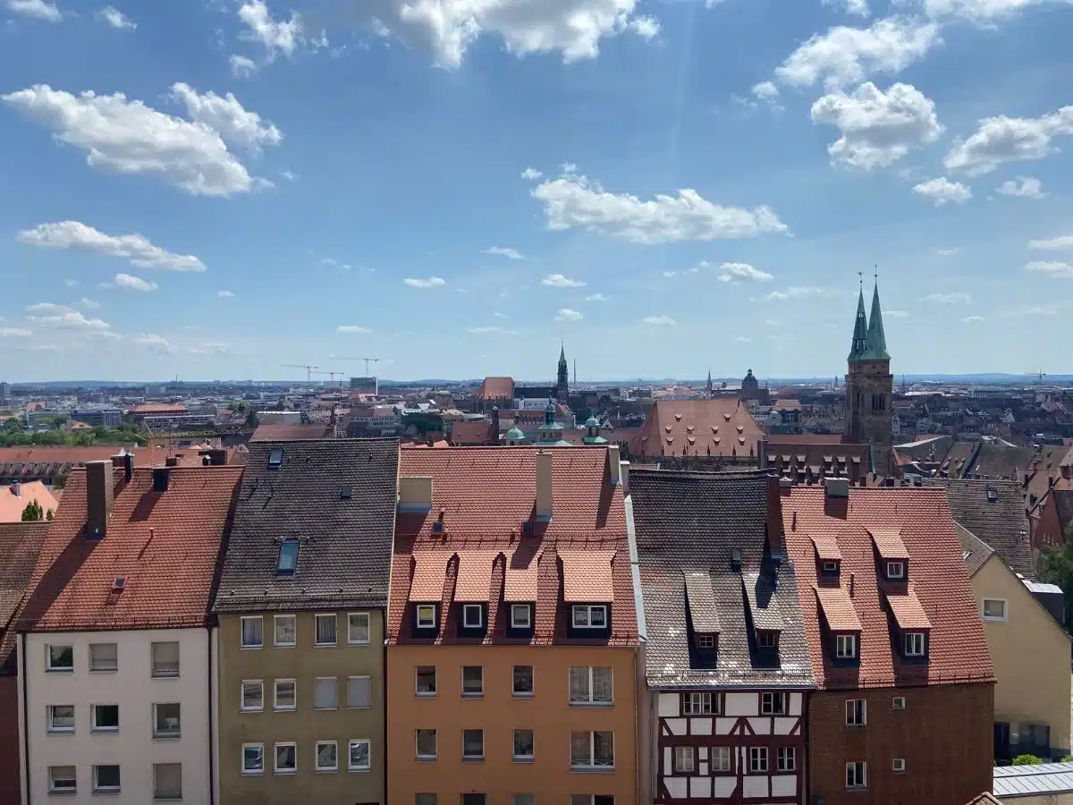 Panoramic view over the rooftops of Nuremberg on a clear day, showcasing the city's blend of traditional and modern architecture. Red-tiled roofs and historic spires rise above the streets, while in the distance, contemporary construction cranes hint at the city's ongoing growth. The blue sky, dotted with fluffy clouds, stretches out, offering a sense of the vast history and vibrant life in Nuremberg.