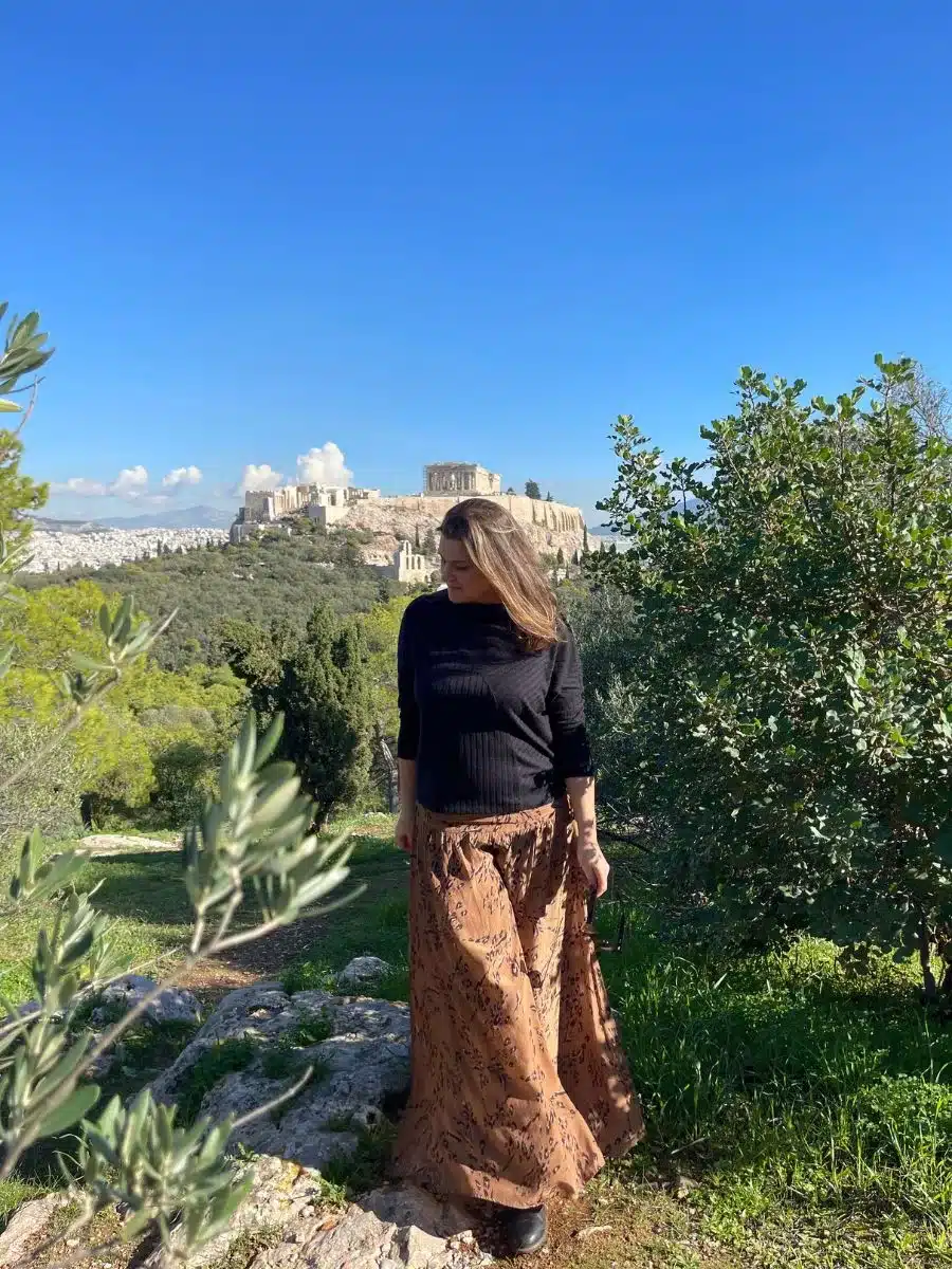 A woman stands contemplatively in a lush green park, with the iconic Parthenon temple poised on the Acropolis in the distance, symbolizing the fusion of natural beauty and historic grandeur in Athens