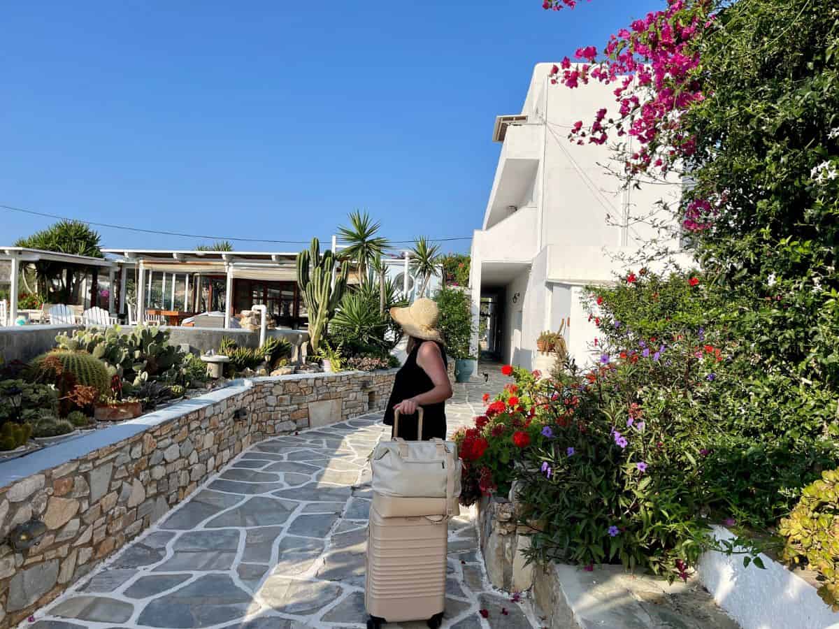A woman and her suitcase manifesting travel to  a greek island village surround by flowers