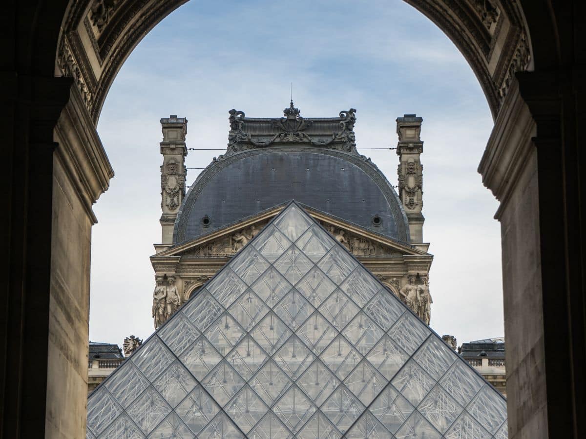 A unique perspective of the iconic Louvre Pyramid with the grand Louvre Palace in the background, framed by an archway, a sight to behold on a weekend in Paris.