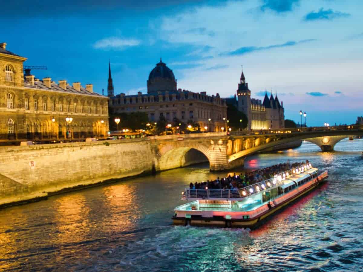 A riverboat cruise glides along the Seine River in Paris during the blue hour, with the city lights reflecting off the water and historic buildings lining the bank, a charming experience for a solo traveler or a weekend in Paris.